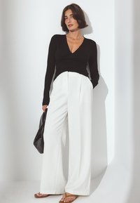 Mujer con cabello corto castaño, vistiendo una blusa de manga larga negra, pantalones anchos blancos, sandalias marrones, sosteniendo un bolso negro, de pie contra una pared blanca.