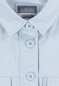 Světle modrá košile na knoflíky vyrobená z denimu. Disponuje špičatým límcem, třemi knoflíky a štítkem s nápisem "SUBLEVEL Medium." Jednoduchá textura.