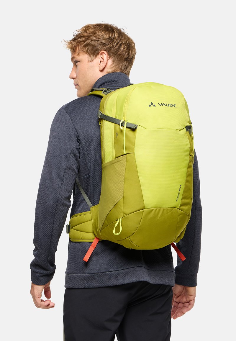 Jeune homme en veste foncée portant un grand sac à dos de randonnée Vaude jaune-vert avec des bretelles rembourrées et une ceinture lombaire, face à gauche.