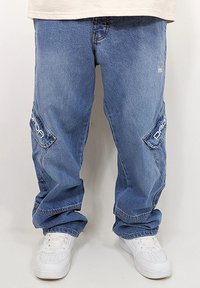 Blue denim cargo pants featuring large side pockets with white logo accents, relaxed fit, and cuffed hems. Paired with white sneakers.