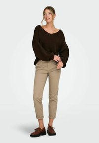 Naine seisab, kandes tumepruuni üle suuruse kampsunit, beežid ülesrullitud pükse ja pruune tikkesussid, vaadates veidi kõrvale.