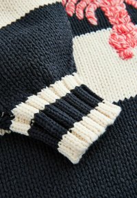 Manche de pull en maille rayée noir et blanc avec une base crème et un motif rouge partiel au-dessus du poignet.