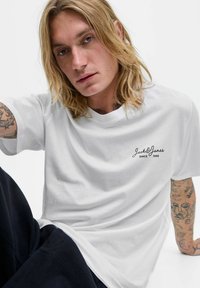 Jaunas vyras su ilgais blondiniais plaukais ir tatuiruotėmis, vilkintis baltą "Jack & Jones" marškinėlį ir juodas kelnes, sėdintis prieš baltą foną.
