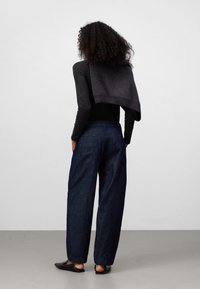 Mörkgrå tröja draperad över en svart långärmad skjorta, parat med lösa mörka denimbyxor och svarta platta skor. Släta texturer och avslappnad design.