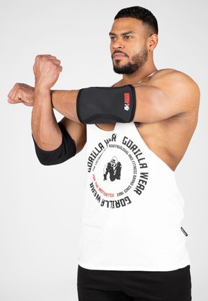 Gespiervde man draagt een wit mouwloos Gorilla Wear-shirt en rekt zijn arm, met zwarte elleboogbeschermers aan beide armen tegen een effen achtergrond.