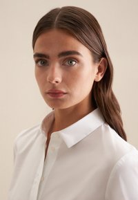 Biała koszula z kołnierzykiem z gładkiej tkaniny, z zapięciem na guziki z przodu i dopasowanym krojem, prezentowana na modelce z długimi brązowymi włosami.