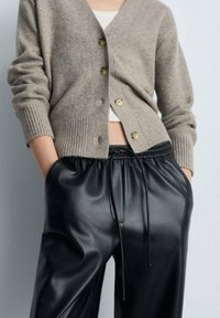 Cardigan en tricot beige avec un col en V et des poignets côtelés, orné de boutons marron. Pantalon noir en simili cuir avec des cordons de serrage et des poches latérales.