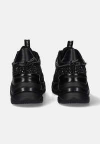 Zapatillas deportivas negras con parte superior de tela texturizada, acentos negros brillantes y suelas de goma curvadas. Incluyen un sistema de cordones seguro con etiquetas.