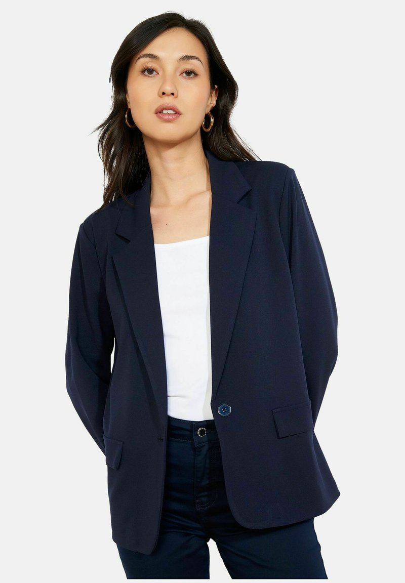 Femme portant un blazer bleu marine foncé sur un haut blanc et un pantalon foncé, debout les mains derrière le dos devant un fond uni.