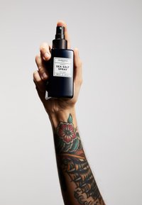 Svart sprayflaska med en matt yta, märkt "Sea Salt Spray." Handen som håller den har en färgglad tatuering. Enkel, neutral bakgrund.