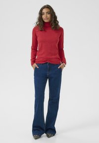 Piros garbó pulóver sötétkék bővülő farmernadrággal párosítva. A pulóver puha tapintású, a farmernadrág oldalsó zsebekkel rendelkezik, és klasszikus szabású.