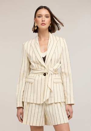 Kvinde iført cremegul pinstripe blazer med matchende shorts og guldbelagte øreringe, stående foran en ensartet beige baggrund.
