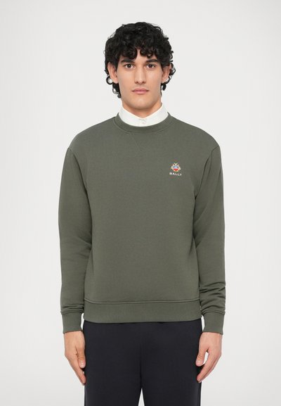 Homme aux cheveux bouclés portant un sweat-shirt Bally vert olive par-dessus une chemise blanche à col, se tenant devant un fond blanc uni.