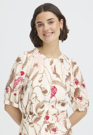 Lühikeste tume juustega naine kannab kreemikat pluusi, millel on pruunid ja punased lillemustrid, naeratab ja poseerib käed selja taga.