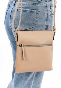 Béžová kožená crossbody taška s jednou zipovou kapsou, která má menší přední kapsu na zip, připevněná k dlouhému popruhu.