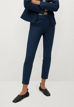 Person iført marineblå slim-fit ankellange bukser, matchende blazer, beige top og sorte flade sko, stående med armene over kors mod en ensfarvet baggrund.