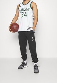 Maillot de basket blanc avec "BUCKS" et le numéro "34", associé à un pantalon de jogging noir portant un logo NBA, des baskets noires Nike et un ballon de basket.