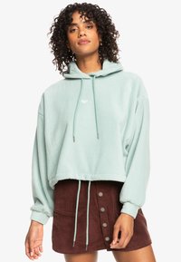 Sudadera corta en verde menta de suave felpa con capucha ajustable, que presenta un pequeño logo y puños elásticos, combinada con una falda de pana marrón.