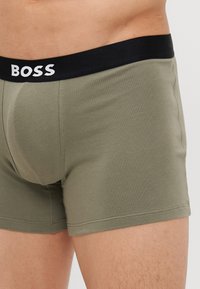 Zeleni pamučni bokserice s crnim pojasom na kojem se nalazi ime brenda "BOSS" napisano bijelim slovima, naglašavajući tijesno pristajanje i glatku teksturu.