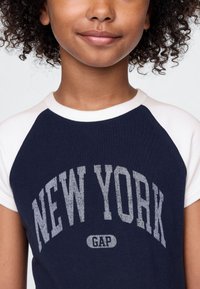 Tamsiai mėlynas ir baltas trumpomis rankovėmis marškinėlis su sidabriniais užrašais „NEW YORK“ ir žemiau esančiu „GAP“ logotipu, pagamintas iš minkšto medvilnės audinio.