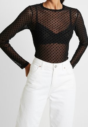 Kvinde iført en sort gennemsigtig langærmet polkaprikket top over en sort bh, kombineret med hvide bukser med høj talje, hænderne hvilende på hofterne.