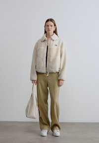 Μπουφάν από φλις εκρού με μεγάλες τσέπες, γιακά και μικρό λογότυπο. Συνδυασμένο με άνετο, ανοιχτόχρωμο χακί παντελόνι και λευκά αθλητικά παπούτσια.