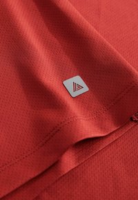 Roter strukturierter Stoff mit einem Netzdesign, versehen mit einem kleinen quadratischen Etikett in Silber mit einem dreieckigen Logo in der unteren Ecke.