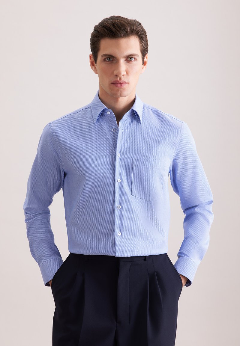 Homme aux cheveux foncés portant une chemise texturée bleu clair rentrée dans un pantalon noir plissé taille haute, mains dans les poches, fond neutre.