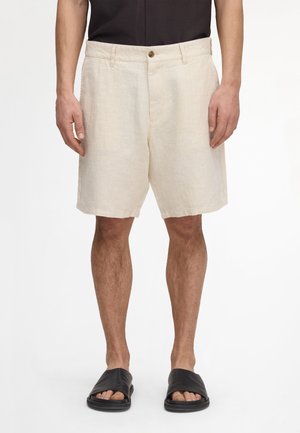 Mann, der beige knielange Shorts, schwarze Sandalen und ein schwarzes Hemd trägt, vor einem weißen Hintergrund steht.