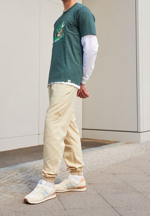 Person trägt ein grünes Grafik-T-Shirt über einem weißen Langarmshirt, beige North Face Jogginghosen und beige New Balance Sneakers.