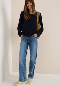 Blouse bleu marine à manches longues avec un col rond, associé à un jean bleu clair à jambes larges. Baskets beiges avec des accents texturés et un sac vert.