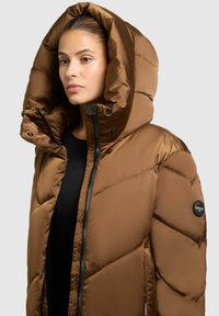 Giacca a vento marrone con un ampio cappuccio, chiusura con zip e bottoni a pressione. Presenta un design trapuntato e una toppa nera con logo sulla manica.