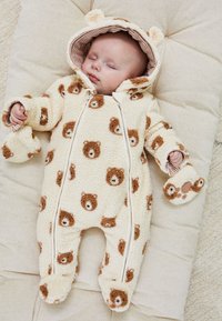 Bébé endormi portant une grenouillère en polaire crème avec des visages d'ours et une capuche avec des oreilles, allongé sur un coussin beige matelassé.