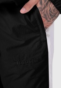 Melnas auduma sporta bikses ar gludu tekstūru, ar sānu kabatu un izliektu "Dropsize" logotipu. Malas apgriezta ar baltu krāsu.