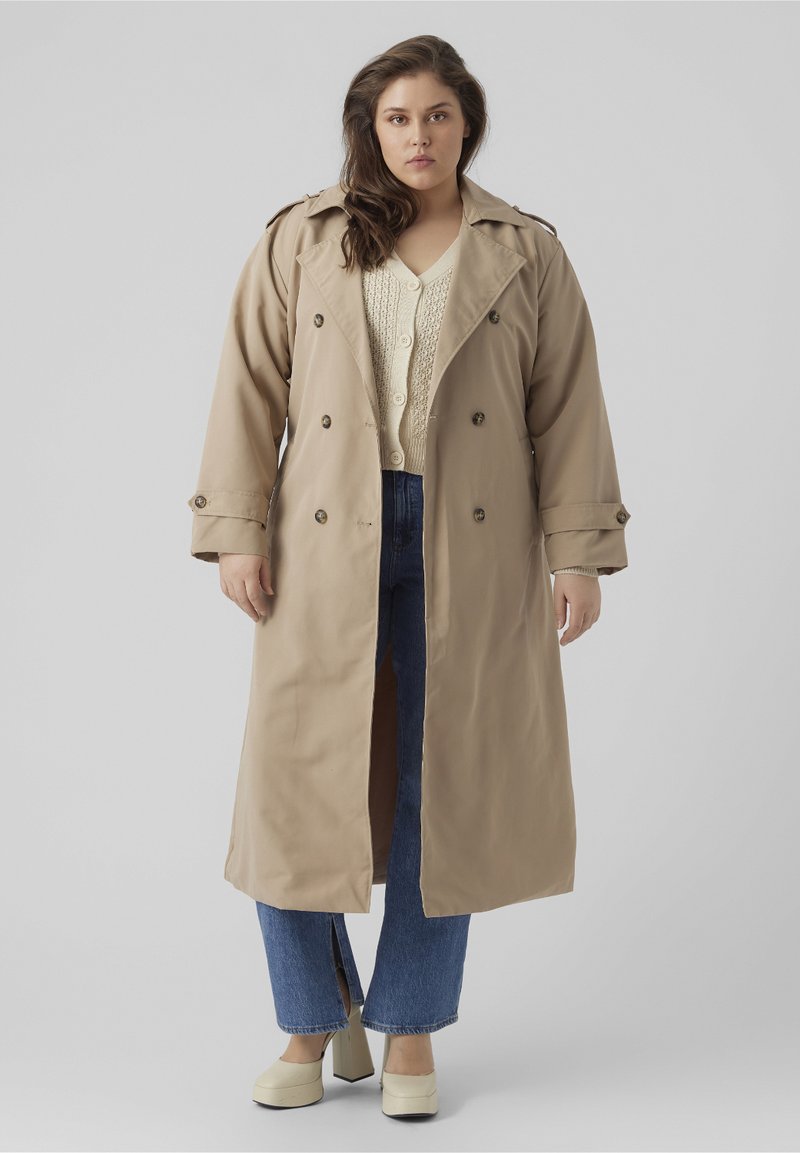 Trench beige con botones de doblebreasted, bolsillos laterales y hombreras. Lleva puesto un suéter crema y vaqueros de cintura alta azules.