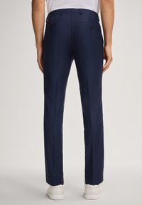 Marineblaue, taillierte Hose mit einer strukturierten Musterung, zwei hinteren Taschen mit schwarzen Knöpfen und einem geraden Bein. Kombiniert mit weißen Sneakers.