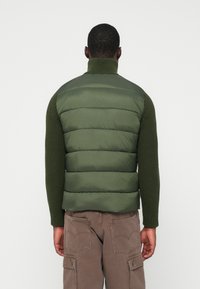 Roheline puffvest kõrge kraega ja ribakudumisega varrukatega. Sile tekstuur horisontaalsete vatitekkide mustriga, vöökoha juures figuuri rõhutav.