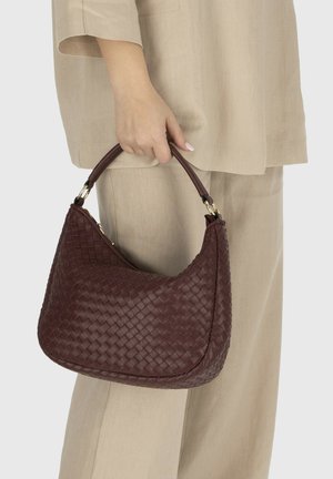 Ręka trzymająca ciemnobrązową plecioną skórzaną torebkę z krótkim uchwytem, w beżowych lnianych spodniach garniturowych i marynarce.