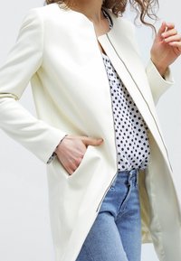 Krämfärgad jacka med rund krage, öppen främre del och sidofickor. Bärs över en blus med prickar och jeans, med en slät yta.