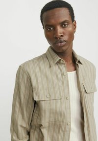 Camicia a maniche corte a righe color marrone chiaro con bottoni sul davanti, due tasche sul petto e una texture morbida. Indossata sopra un top in cotone bianco a coste.