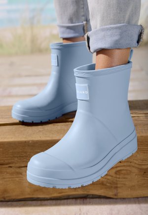 Person wearing light blue rain boots with rolled-up jeans standing on wooden surface near a blurred outdoor background.