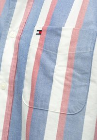 Ριγέ πουκάμισο με οριζόντιες κόκκινες, μπλε και λευκές ρίγες, με τσέπη στο αριστερό στήθος και ένα μικρό κεντημένο λογότυπο. Κατασκευασμένο από ελαφρύ ύφασμα.