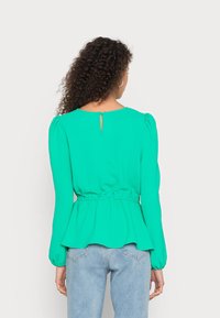 Top verde a maniche lunghe con vita arricciata, dettaglio a occhio di bue sul retro e maniche ampie, realizzato in un tessuto leggero. Abbinato a jeans chiari.