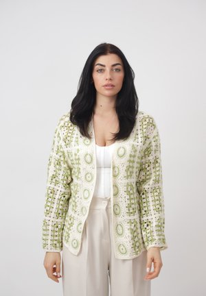 Femme aux longs cheveux bruns portant un cardigan vert et blanc en crochet par-dessus un haut blanc et un pantalon beige taille haute, debout devant un fond uni.