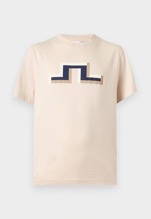 Muž s béžovou čiapkou, béžovým tričkom s logom, khaki šortkami, čiernymi ponožkami a teniskami, ktorý drží čiernu tašku na jednotnom pozadí.