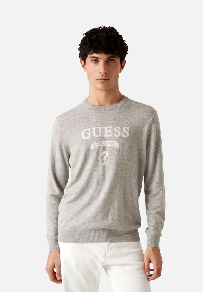Suéter gris hecho de una tela suave, con cuello redondo, mangas largas y un diseño frontal con el logo "GUESS" y el año "1981".
