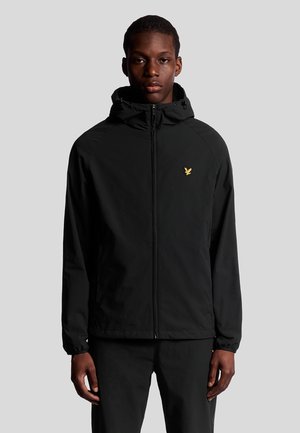 Giacca nera con zip e cappuccio, realizzata in materiale leggero. Presenta un logo dorato sul lato sinistro del petto, polsini elastici e un design semplice.