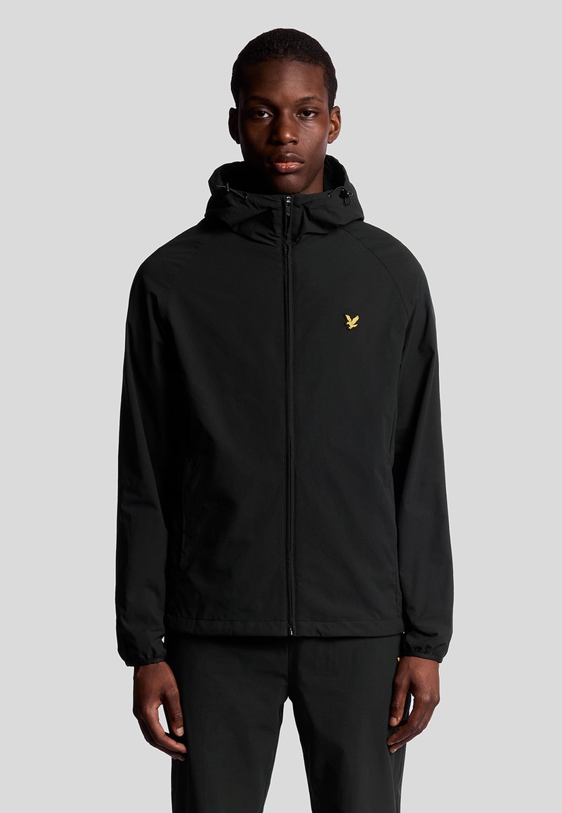 Veste noire à fermeture éclair avec capuche, fabriquée en matériau léger. Présente un logo doré sur la poitrine gauche, des poignets élastiques et un design simple.