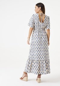 Maxikleid in Creme mit blauem auge-förmigem Muster. Merkmale sind kurze Puffärmel, eine geraffte Taille und ein offener Rücken mit Bindeschluss. Volantsaum.