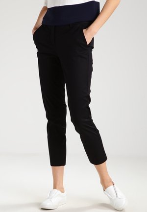 Personne portant un pantalon noir slim court, des baskets blanches sans lacets, et un haut bleu marine et blanc, debout les mains dans les poches.